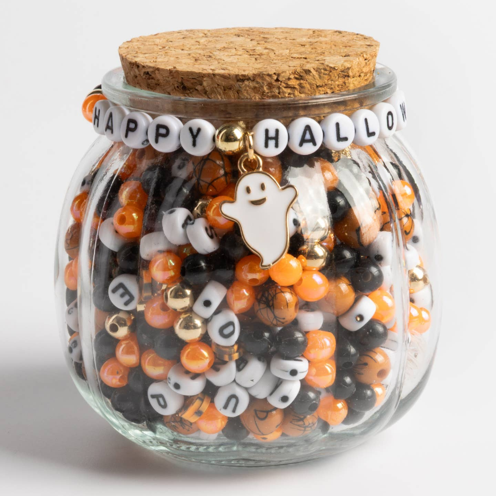 Pumpkin-shaped glass jar with cork lid containing orange and black beads and  white letter beads. The bracelet on the front reads "happy halloween" with a ghost charm.
