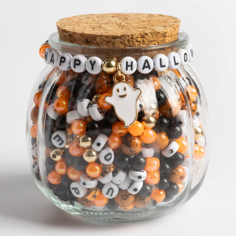Pumpkin-shaped glass jar with cork lid containing orange and black beads and  white letter beads. The bracelet on the front reads "happy halloween" with a ghost charm.