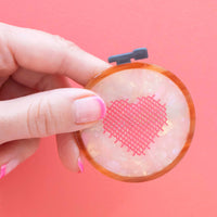 Hand holding a small round hair clip with a pink heart design against a pink background