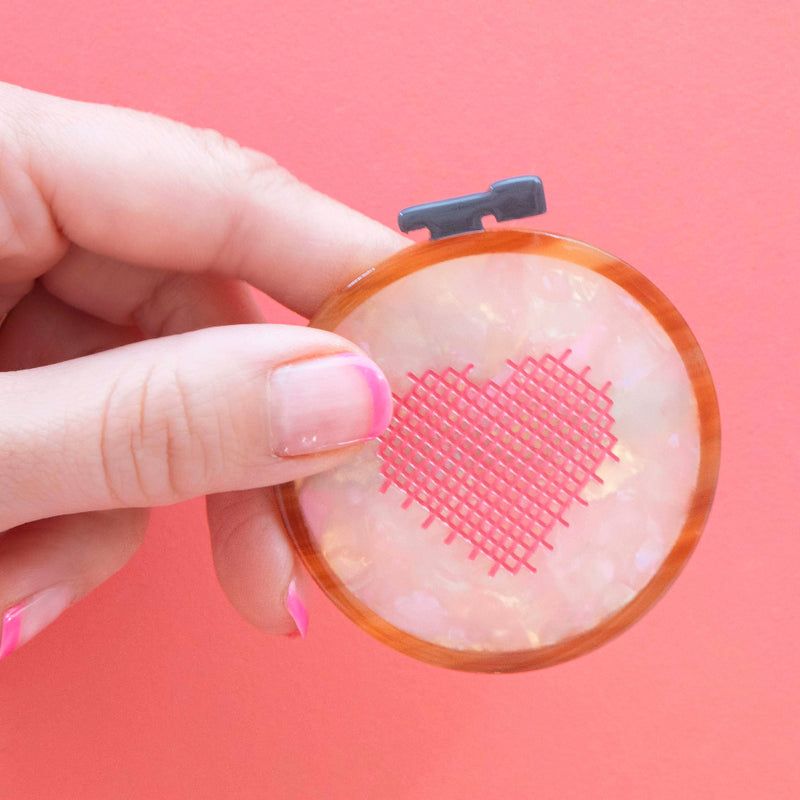Hand holding a small round hair clip with a pink heart design against a pink background
