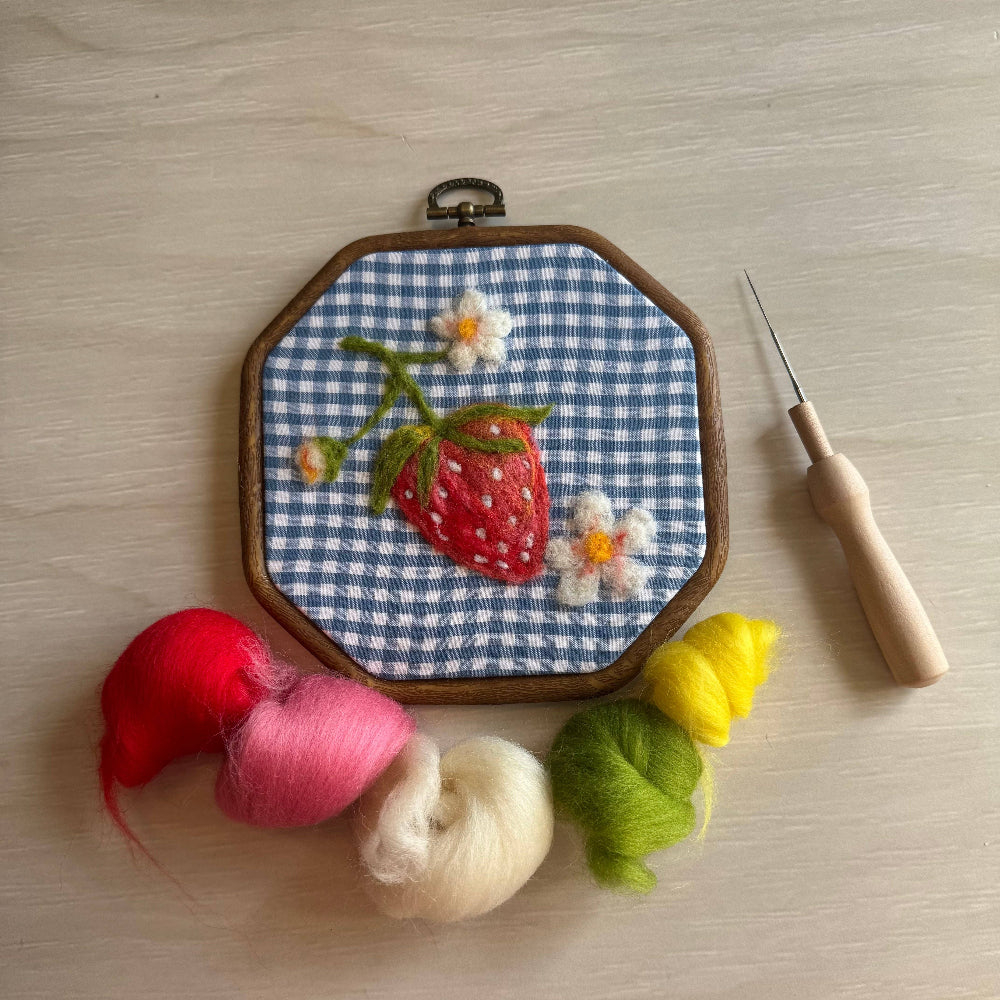 Embroidery hoop with strawberry design on checkered fabric, surrounded by yarn and a needle on a wooden surface.