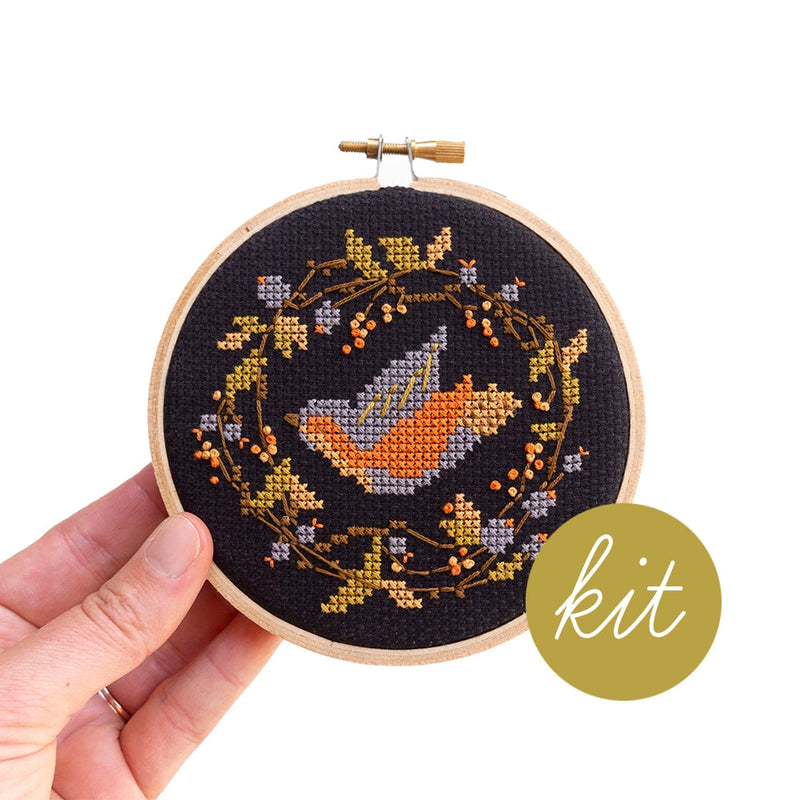 Hand holding an embroidery hoop with blue, orange, and yellow bird cross stitched on a black background and surrounded by a leafy garland.