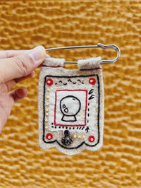 Handmade felt pin with crystal ball design held by a hand on a textured brown background