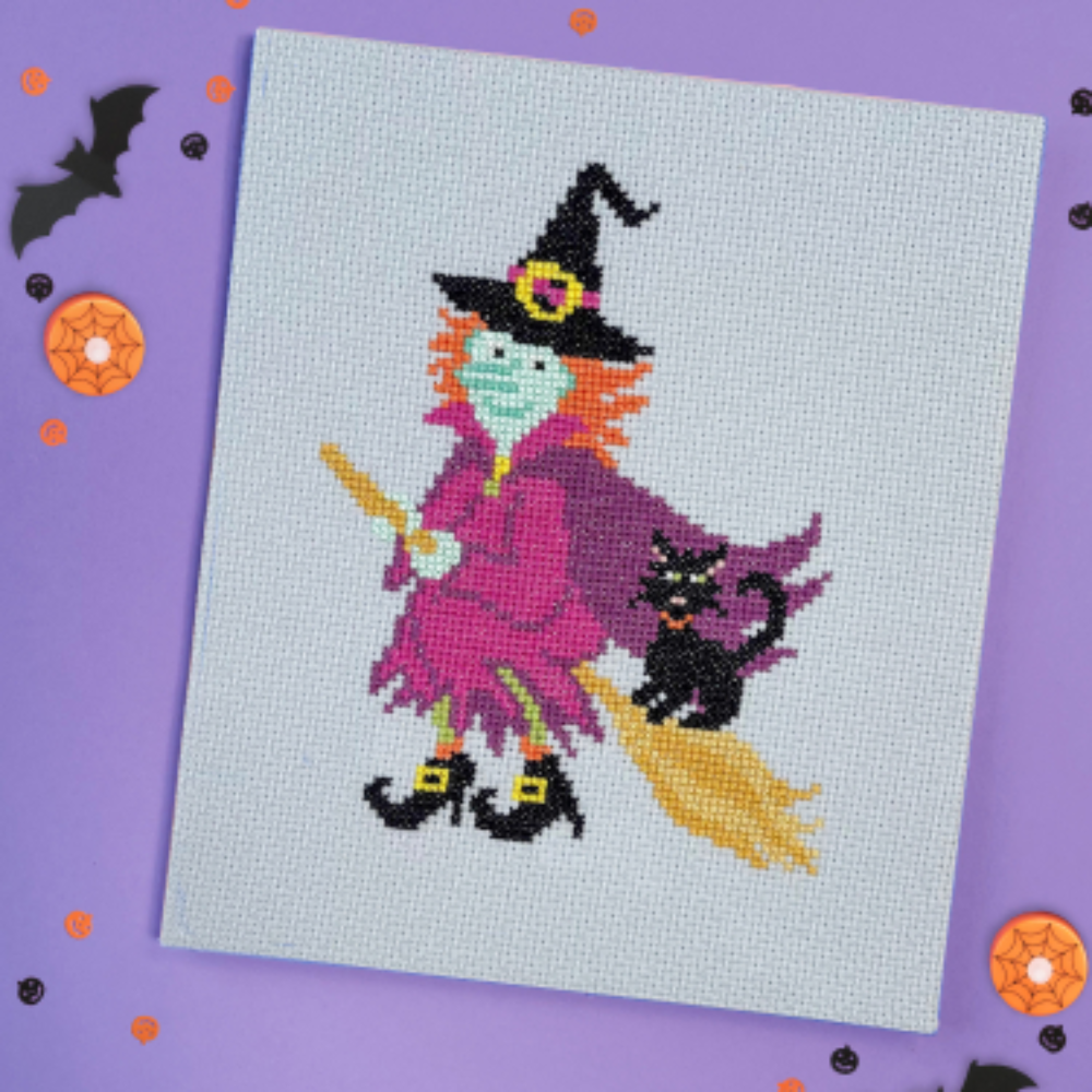 Cross stitch canvas showing a witch on a broom with orange hair, purple clothes, and a black cat. Shown on a purple halloween themed background.