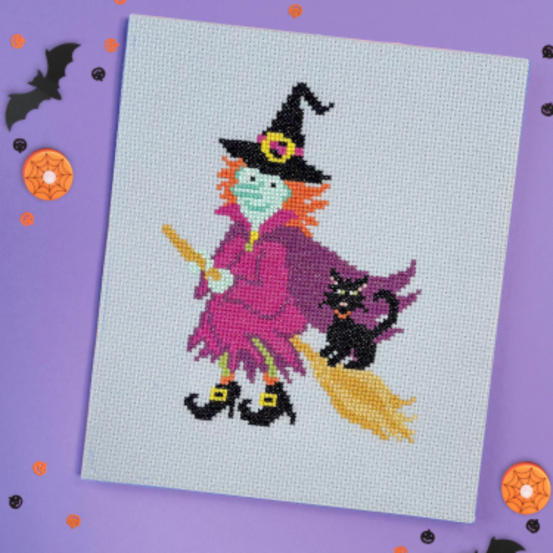 Cross stitch canvas showing a witch on a broom with orange hair, purple clothes, and a black cat. Shown on a purple halloween themed background.