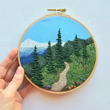 An embroidery kit that depicts a hiking trail with mountain peeks in the background, pine trees, and wildflowers