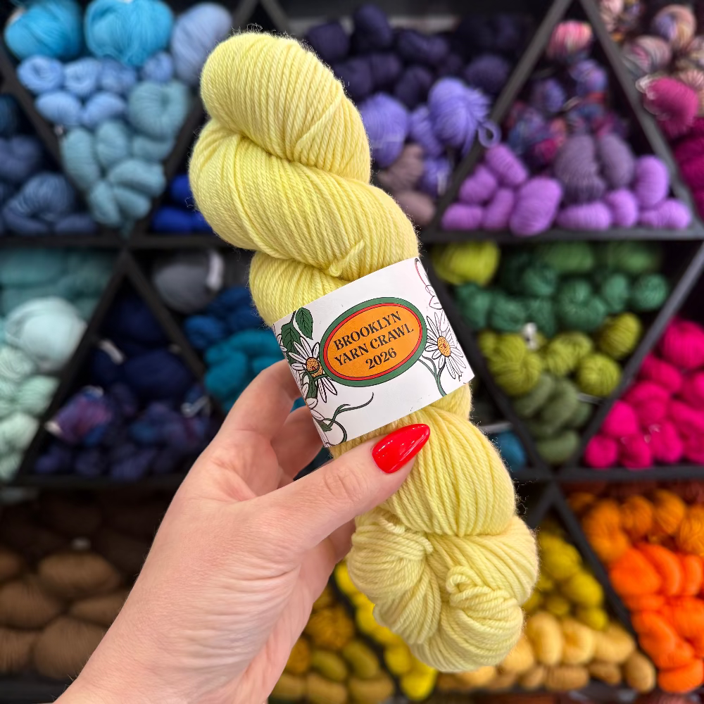 Hand holding a skein of yellow yarn with a label in front of a colorful yarn display.