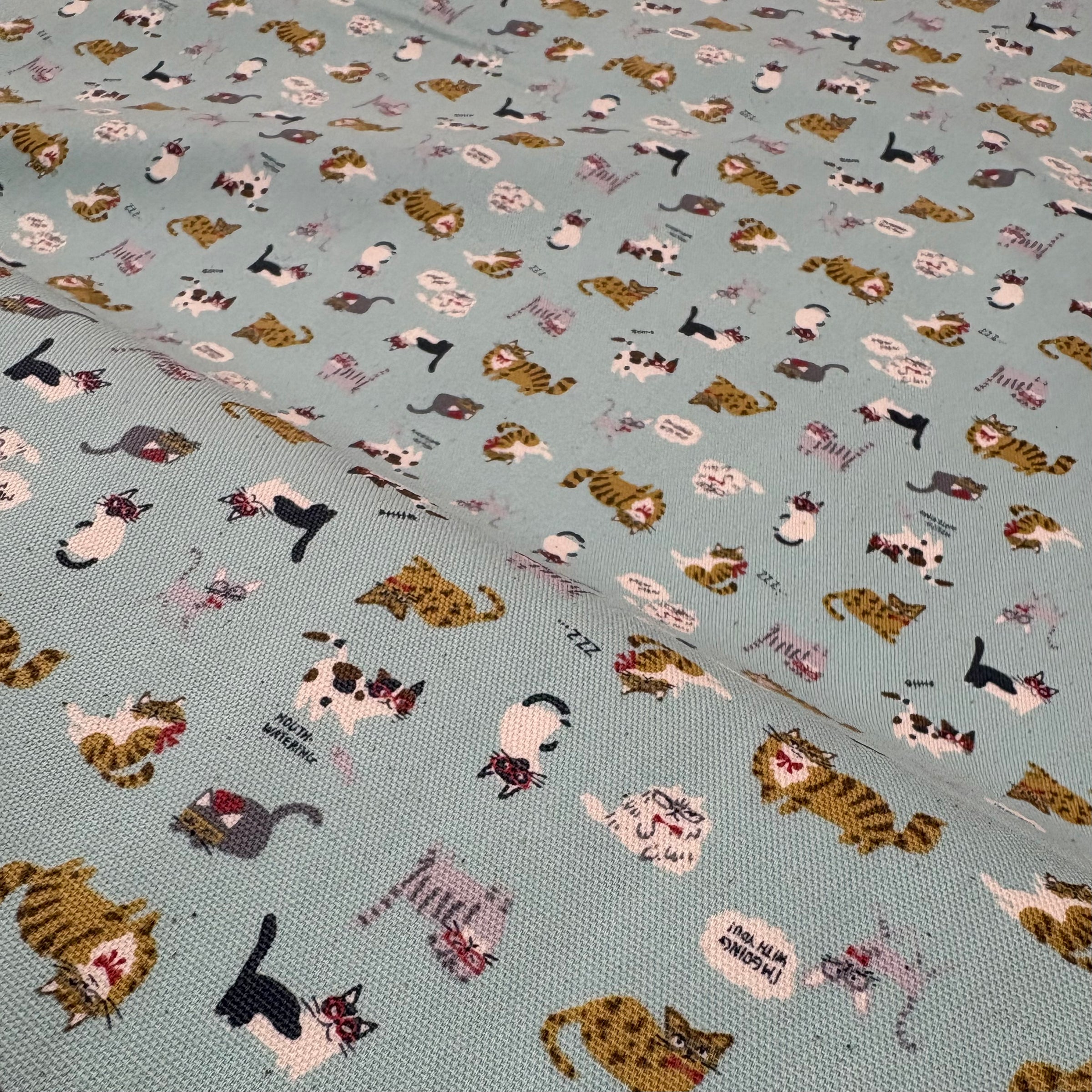 Close up of a bolt of light blue fabric featuring illustrations of cats wearing glasses and bowties.