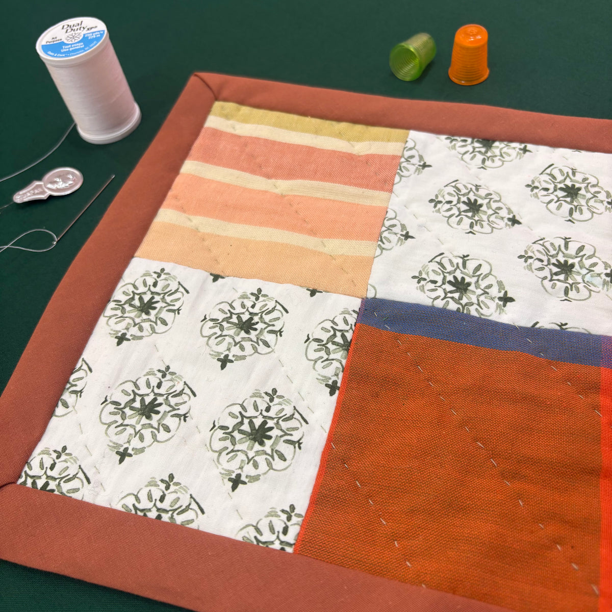 A colorful miniature patchwork quilt on a green background, along with two thimbles, a spool of thread, a sewing needle, and a needle threader