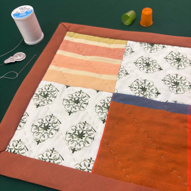 A colorful miniature patchwork quilt on a green background, along with two thimbles, a spool of thread, a sewing needle, and a needle threader