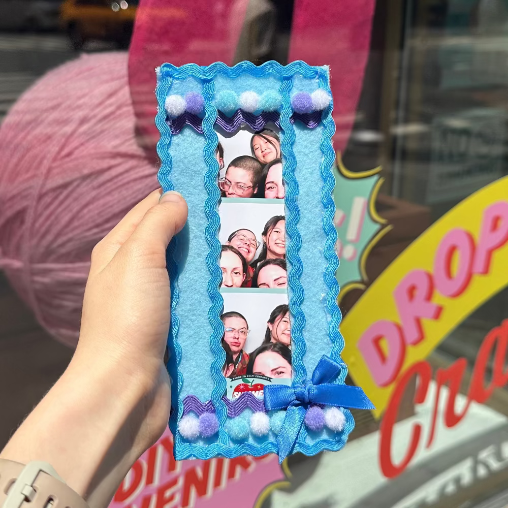 A hand holds a decorative blue photo frame with scalloped edges, purple pom-pom accents, and a small blue ribbon bow, photographed in front of a craft shop window with yarn balls and a 'Drop-In Crafts' sign visible in the background.