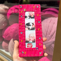 A hand holds a bright pink handmade felt photo frame with embroidered details and rhinestones, featuring three photo slots with a dog portrait. The background shows a shop window with pink yarn balls.