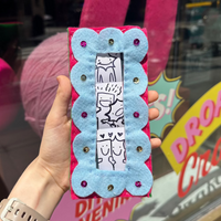 A hand holds a handmade felt pocket frame in front of a craft shop window. The frame features a light blue scalloped border and a hot pink center