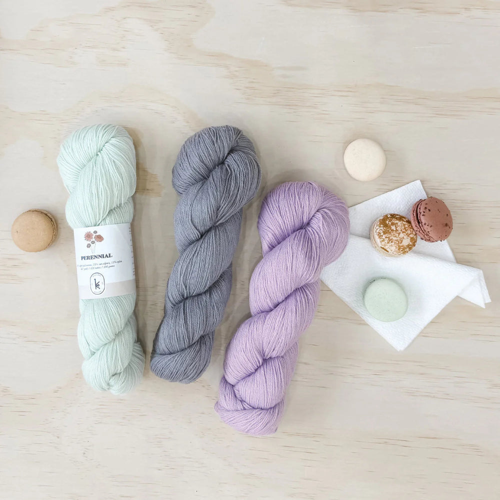 Three skeins of yarn in light green, gray, and purple on a wooden surface with small macarons.
