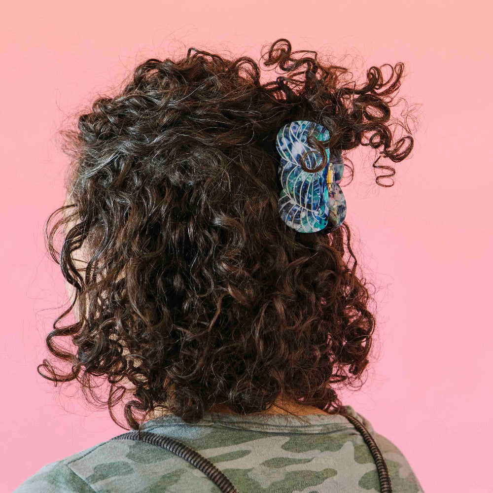 Person with curly hair wearing a colorful yarn skein hair clip on a pink background