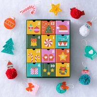A colorful Christmas advent calendar with 12 numbered compartments surrounded by festive handmade ornaments on a white snowy background.