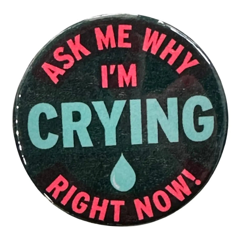 A black button against a white background. Text on the button reads "Ask Me Why I'm Crying Right Now" in red text with the word crying and a tear drop in the center in blue. The surrounding text is in bright pink.