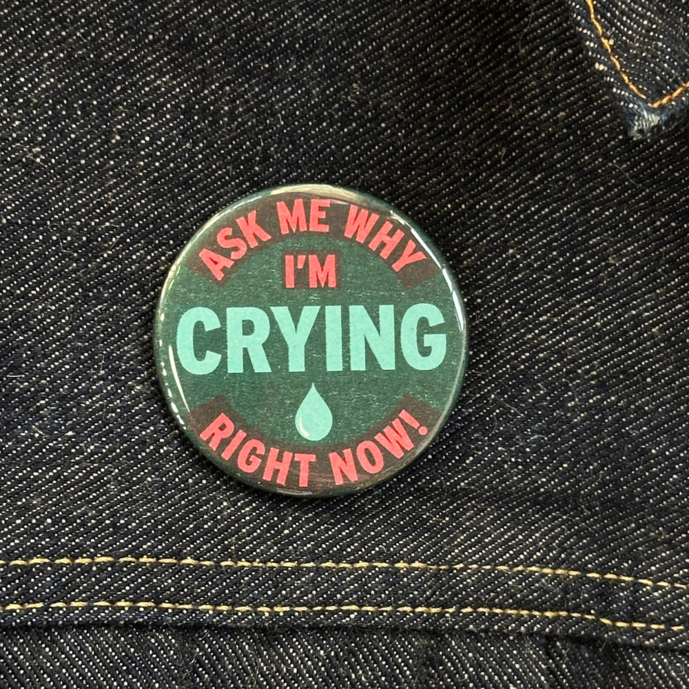 A button is placed on a denim jacket near the collar for size comparison. Text on the button reads "Ask Me Why I'm Crying Right Now" in red text with the word crying and a tear drop in the center in blue. The surrounding text is in bright pink. 