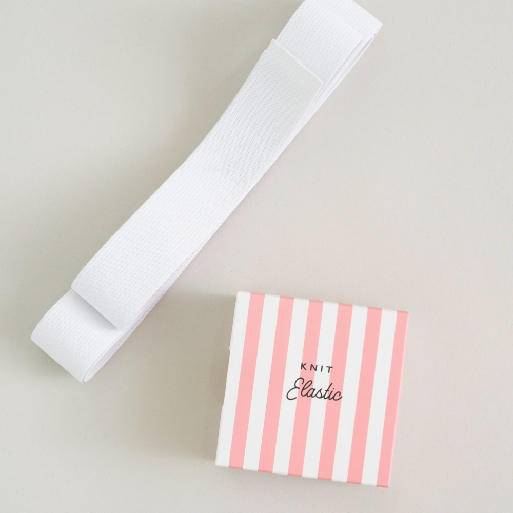 A white knit elastic band is laid diagonally next to its pink and white striped packaging box with 'KNIT Elastic' printed on it, set against a neutral light gray background.