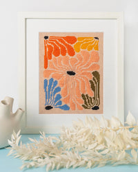 Framed needlepoint art with orange, blue, pink, and green flowers on a light blue surface with a white background