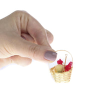 A hand holding a miniature wicker basket containing yarn and knitting needles.