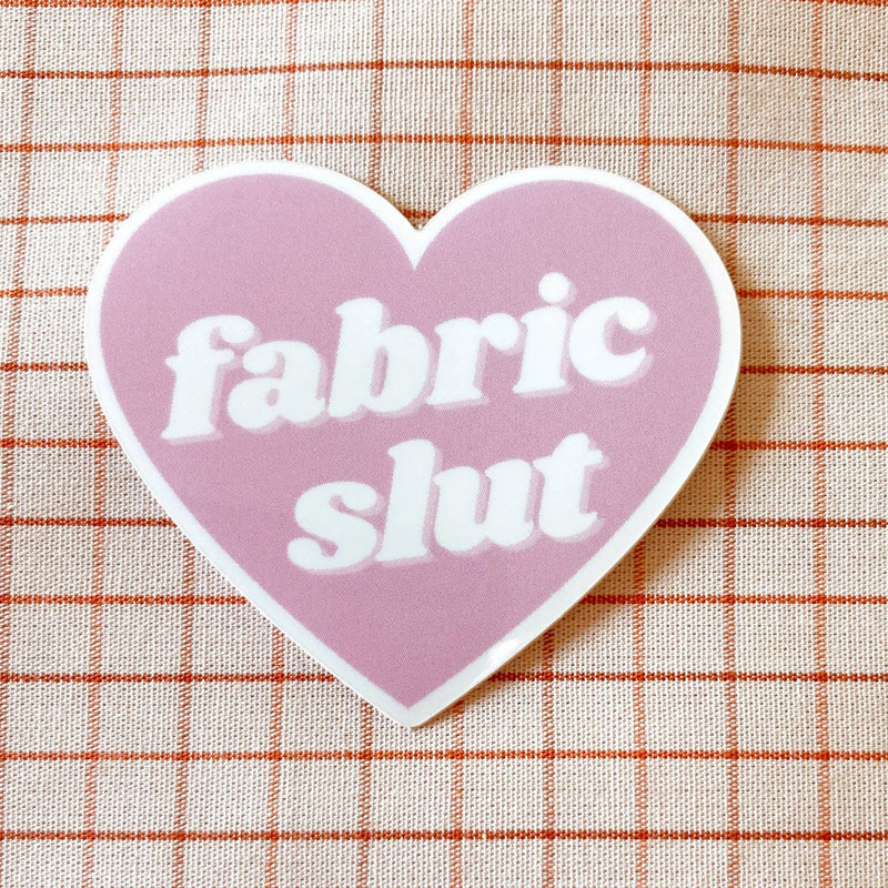 Heart-shaped sticker with 'fabric slut' text on a checkered fabric background