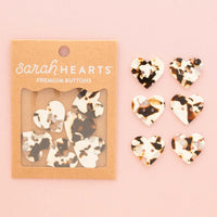 Sarah Hearts premium heart-shaped buttons in a tortoiseshell pattern, shown in their kraft paper packaging alongside six loose buttons on a soft pink background.