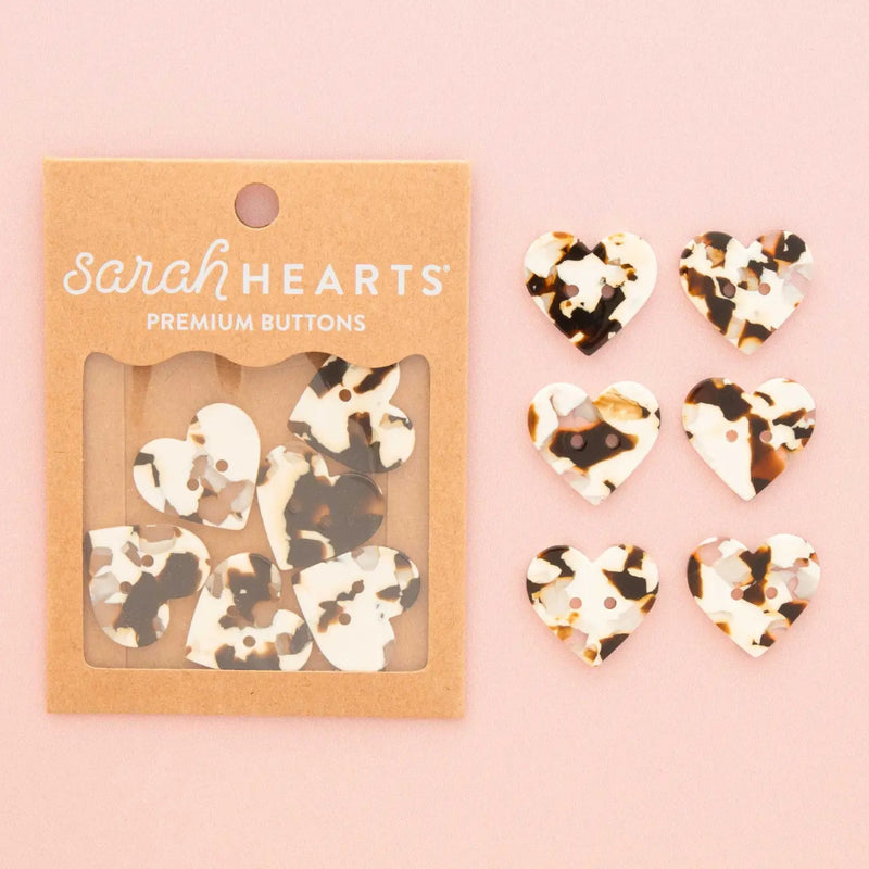 Sarah Hearts premium heart-shaped buttons in a tortoiseshell pattern, shown in their kraft paper packaging alongside six loose buttons on a soft pink background.