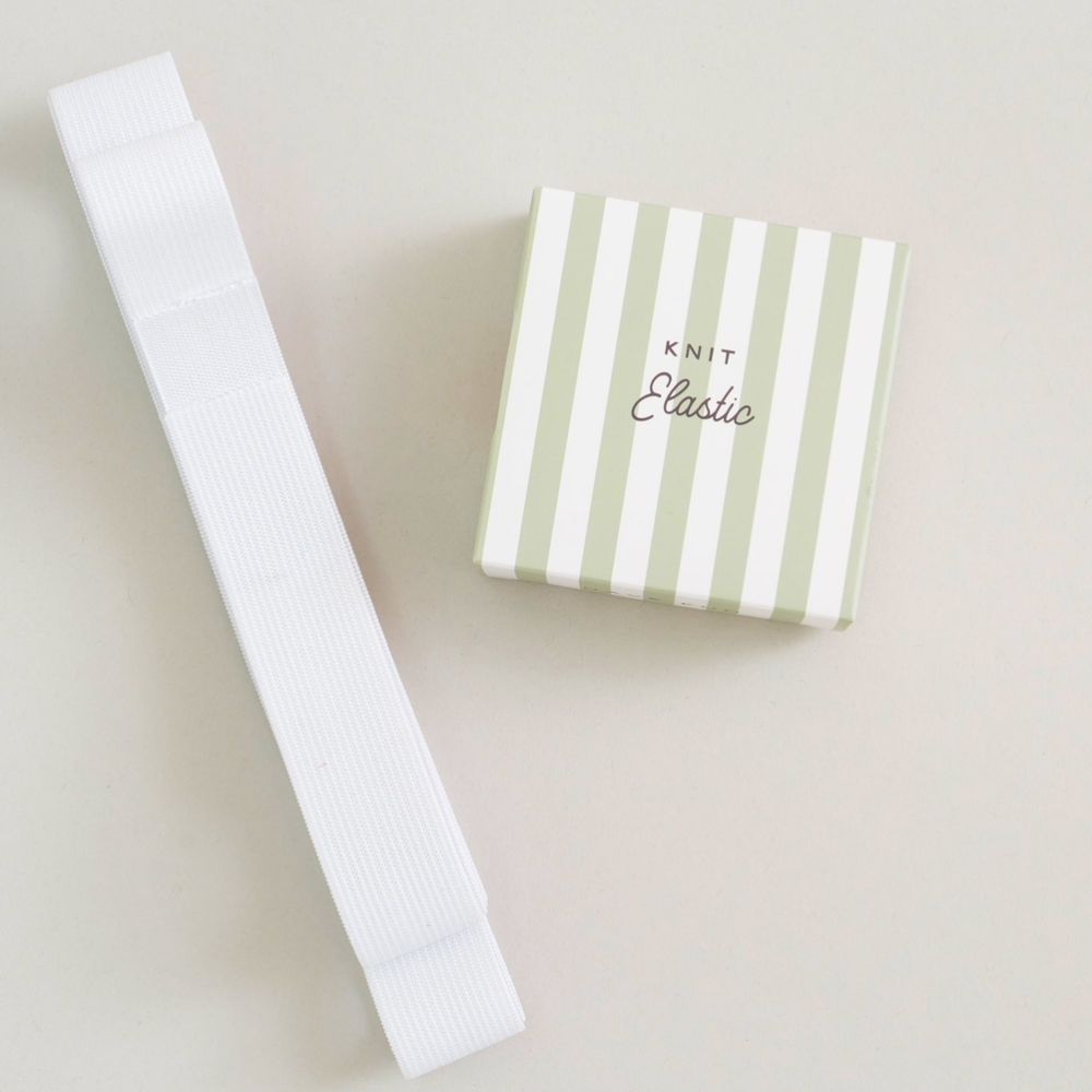 A white knit elastic laid diagonally next to its light green and white striped gift box labeled 'KNIT Classic' on a warm off-white background.