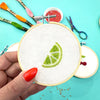Embroidery hoop with green embroidered design held by a hand on a turquoise background with embroidery tools.