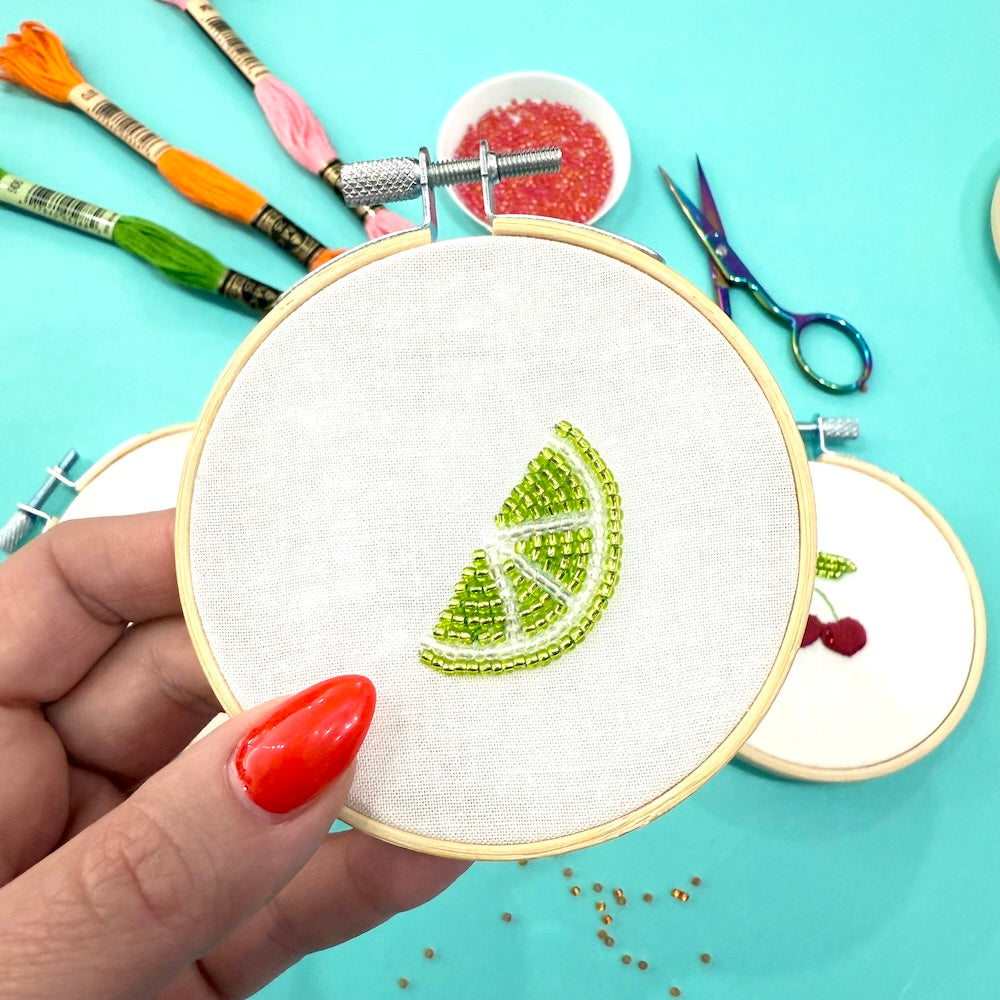 Embroidery hoop with green embroidered design held by a hand on a turquoise background with embroidery tools.