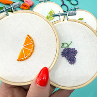 Embroidery hoops with fruit designs held by a hand with red nail polish on a turquoise background.