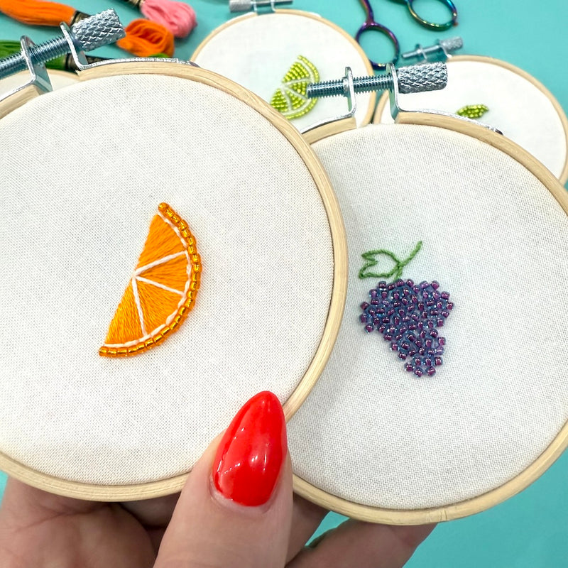 Embroidery hoops with fruit designs held by a hand with red nail polish on a turquoise background.