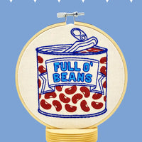 A circular embroidery hoop mounted on a stand, featuring an embroidered design of a can of beans labeled 'FULL O' BEANS' in blue on an off-white fabric background, with red beans scattered below. The hoop is photographed against a light blue background with small white geometric shapes at the top.