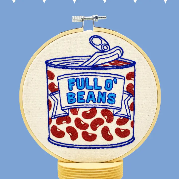 A circular embroidery hoop mounted on a stand, featuring an embroidered design of a can of beans labeled 'FULL O' BEANS' in blue on an off-white fabric background, with red beans scattered below. The hoop is photographed against a light blue background with small white geometric shapes at the top.