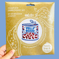 A hand holding a paper package for a complete embroidery kit. The package features white line art of hands holding a needle and a central illustration of a can of 'Full O' Beans' filled with red beans.