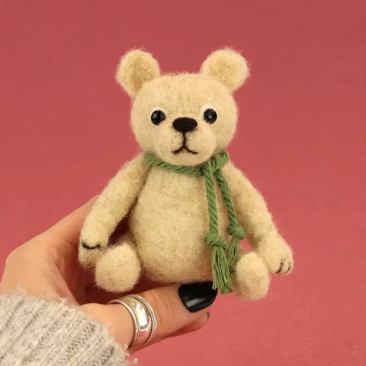 Small beige teddy bear with a green scarf held by a person against a pink background