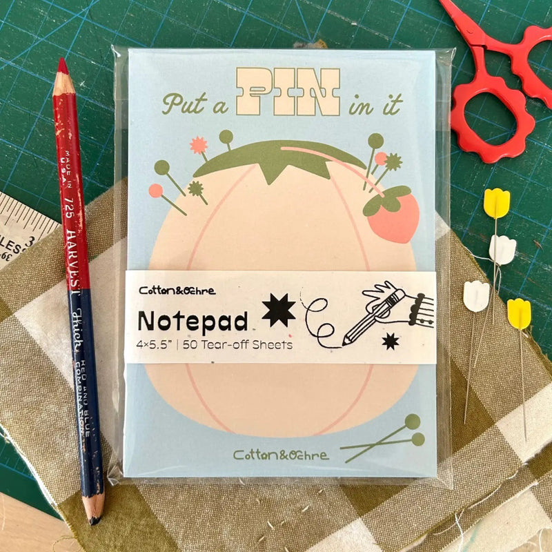 Notepad with a pin-themed design on a crafting surface with a pencil and scissors.