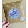 Hand holding a complete embroidery kit packaging with a heron design on a beige background