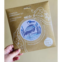Hand holding a complete embroidery kit packaging with a heron design on a beige background