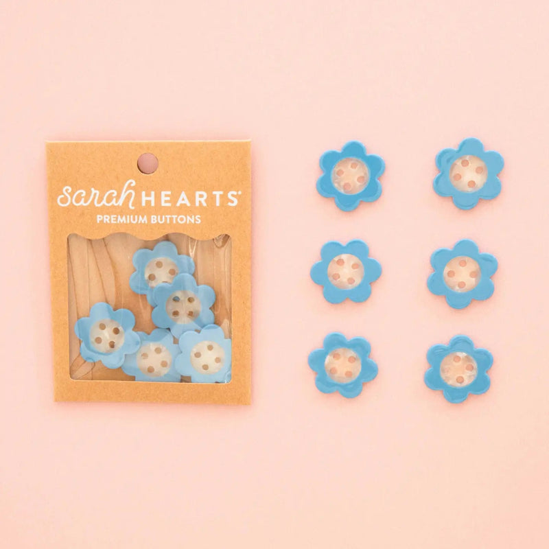 Sarah Hearts flower-shaped buttons in light blue with white centers, shown both inside their kraft paper packaging and arranged in two lines against a soft pink background.