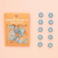 Sarah Hearts daisy shaped buttons in light blue and white, shown in their kraft paper packaging on the left and arranged in two lines on the right against a soft peach background.
