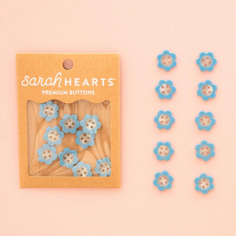 Sarah Hearts daisy shaped buttons in light blue and white, shown in their kraft paper packaging on the left and arranged in two lines on the right against a soft peach background.