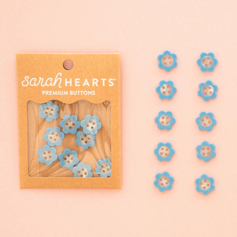 Sarah Hearts daisy shaped buttons in light blue and white, shown in their kraft paper packaging on the left and arranged in two lines on the right against a soft peach background.