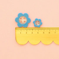 Two blue daisy-shaped buttons (one 1" button and one 5/8" button) with pearl white centers, shown above a yellow scalloped edge ruler on a pale peach background.