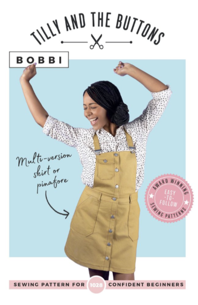 Model wearing a tan overall skirt over a white and black polka dot long sleeve shirt while dancing with hands in the air. She's placed on a light blue and white background with Tilly and the Buttons branding and text.