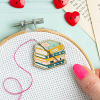 Embroidery hoop with colorful stack of books needle minder and pink thread on a light blue background.