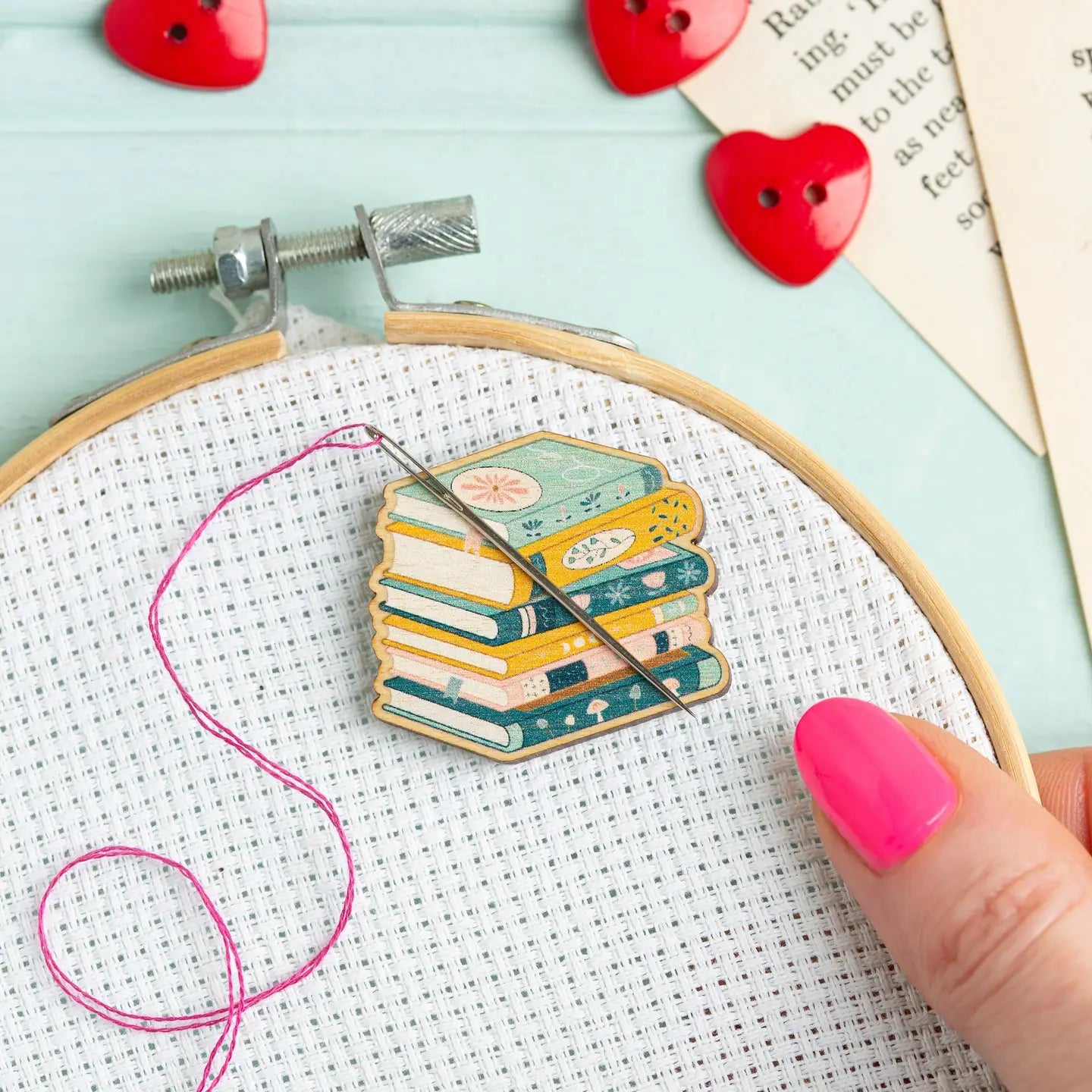 Embroidery hoop with colorful stack of books needle minder and pink thread on a light blue background.