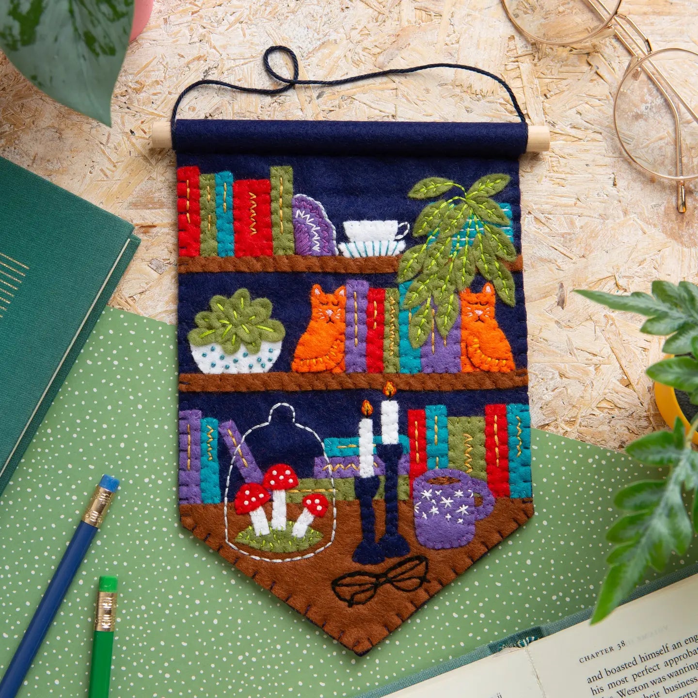 Colorful felt pennant featuring bookshelves, cats, plants, candles, mushrooms, glasses, and a coffee mug laying on a wooden table.