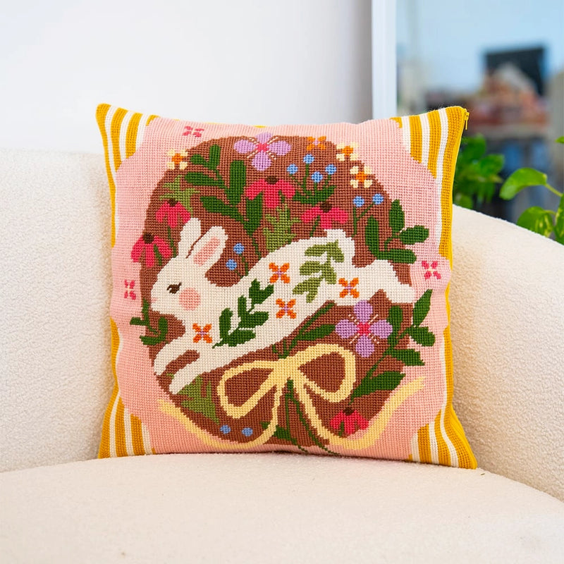 A needlepoint cushion with a floral design featuring a white rabbit sits on a couch.